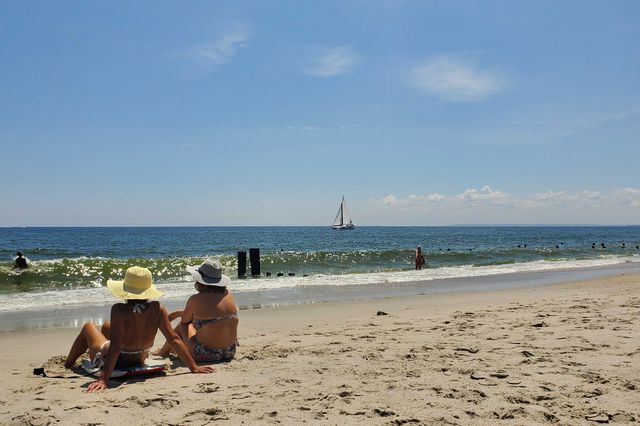 Two women sit on the beach at Jacob Riis park on July 25th as a sailboat with white sails glides by.